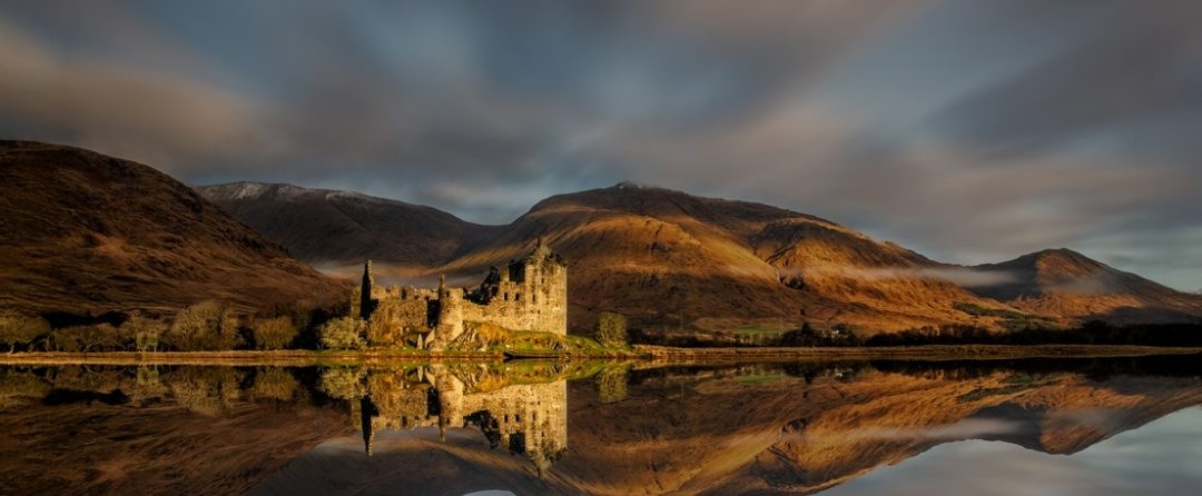 Castle ruins reflected in a Scottish loch beneath golden hills